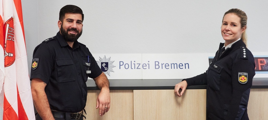 Dunkelhaariger Polizist steht neben blonder Polizistin vor einem Schild mit der Aufschrift "Polizei Bremen"